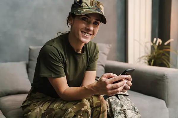 Smiling soldier in military uniform and hat sitting on sofa while using mobile phone