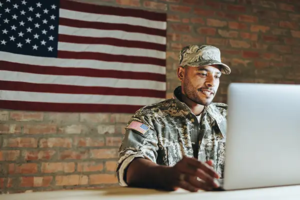 Soldier in US military uniform using laptop with American flag in background on wall